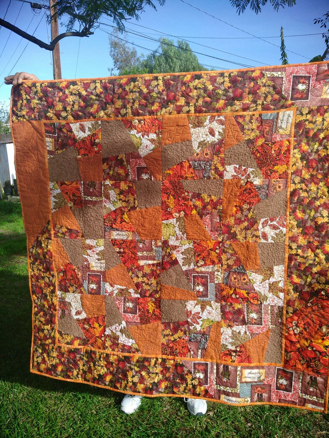 Orange and brown fall crazy quilt held up outdoors. Only hands and shoes are visible.