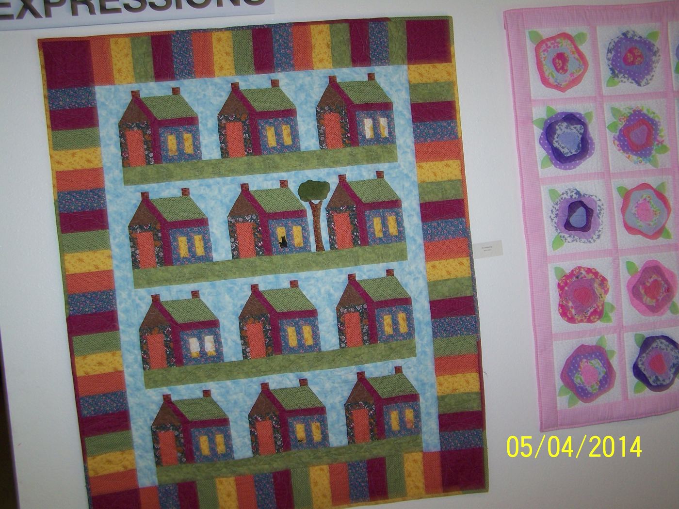 House-block quilt with a striped border, hung on a show wall beside a pink rose quilt.