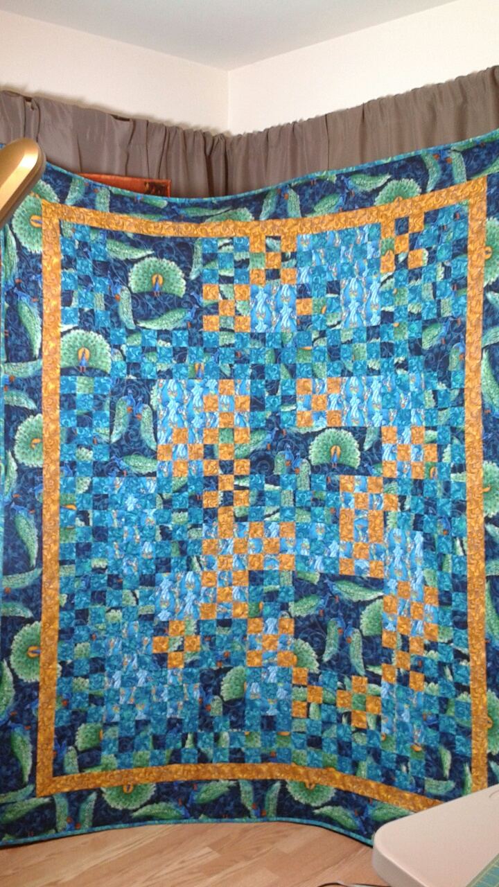 Blue and gold peacock-print quilt hung in a room, with a sewing machine in the foreground.