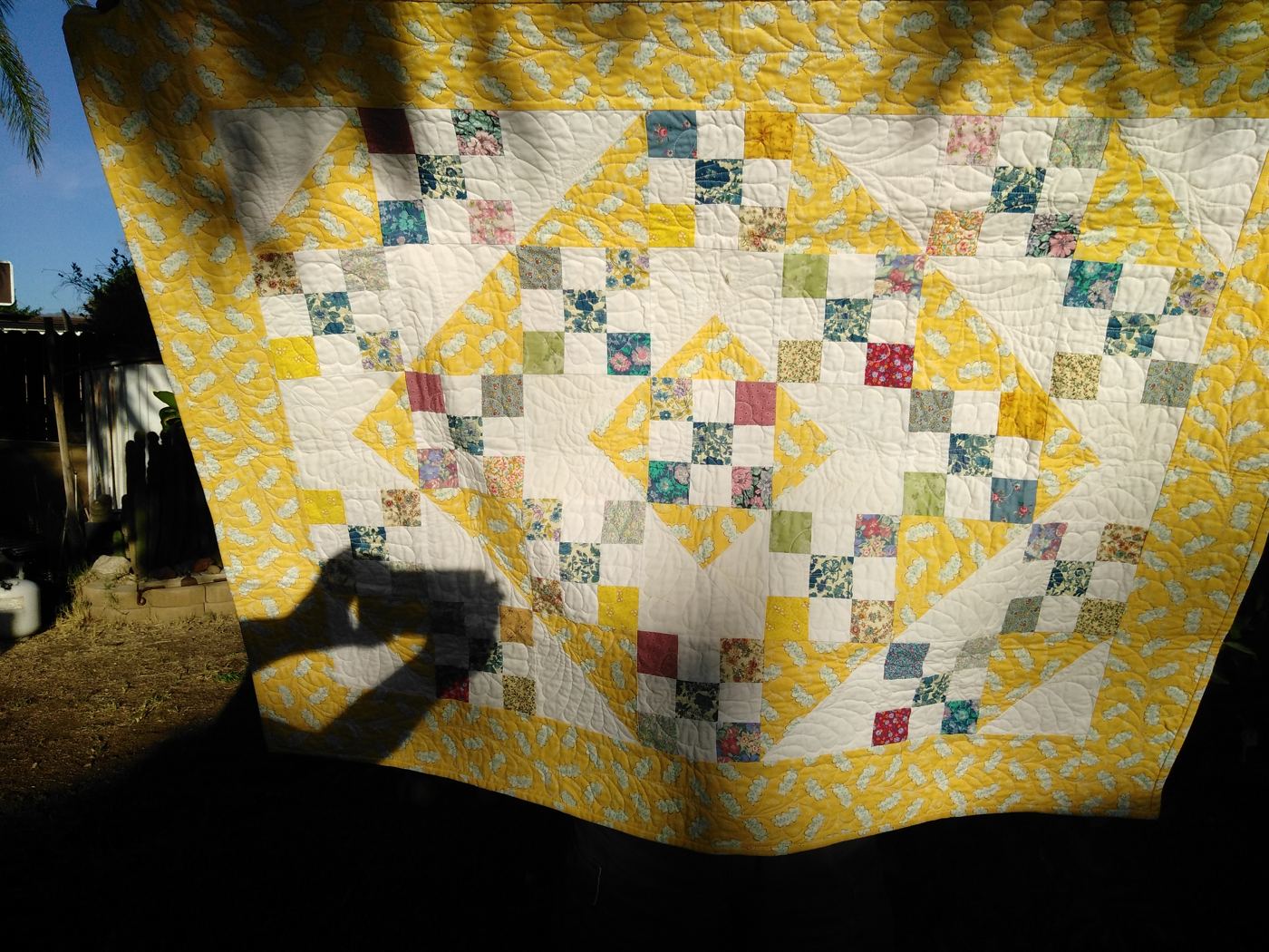 Yellow scrap quilt with nine-patch blocks, hanging outdoors.