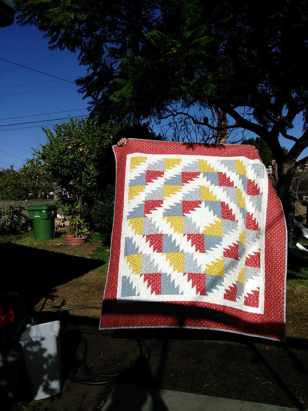 A red, yellow, and blue sawtooth quilt held up in a yard under a tree.